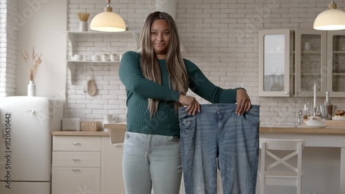 Cheerful attractive plus size African American woman in casual clothes showing oversized jeans, celebrating successful weight loss diet plan, expressing happiness and excitement at home.