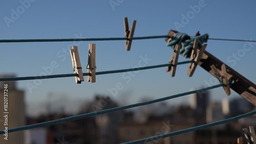 Clothespins hanging on a clothesline against a clear blue sky. The background features blurred buildings, creating an urban atmosphere.