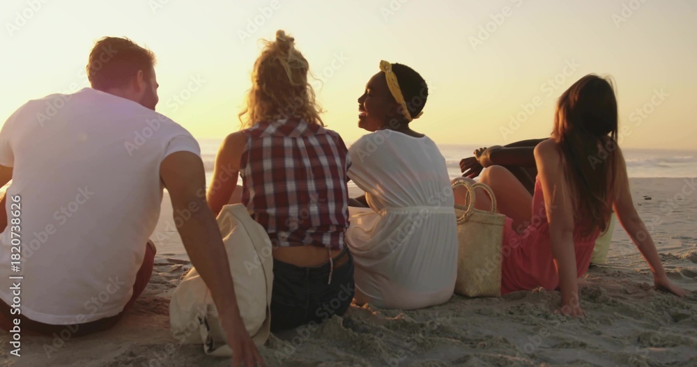 Fototapeta premium Sitting five friends in summer clothes facing ocean at beach, with straw tote and canvas bag
