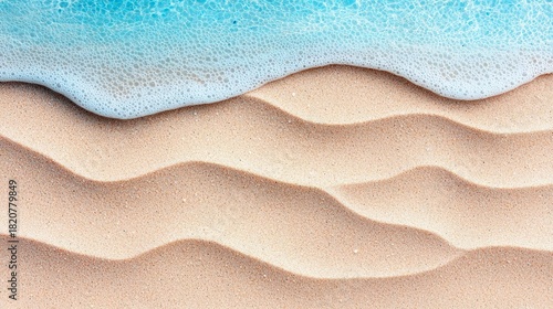 Fototapeta Naklejka Na Ścianę i Meble -  A close-up, top-down view of a sandy beach with gentle dunes and ripples, where a soft wave with white foam is washing over the sand. The water is a clear, brig