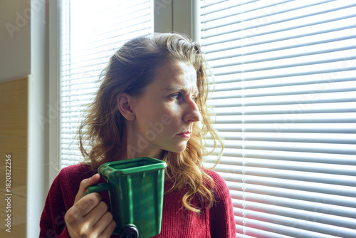 A woman stands by a window holding a green trash-bin mug and looking displeased. Moment of morning frustration in natural light. Concepts of irritation, low-energy routine, and emotional realism.