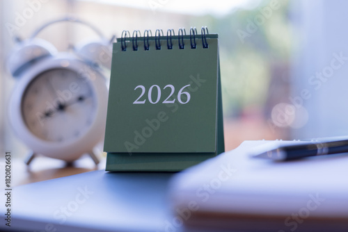 Close-up of green 2026 desk calendar with blurred clock and notebook in background, ideal for new year planning, time management, and productivity visuals.
