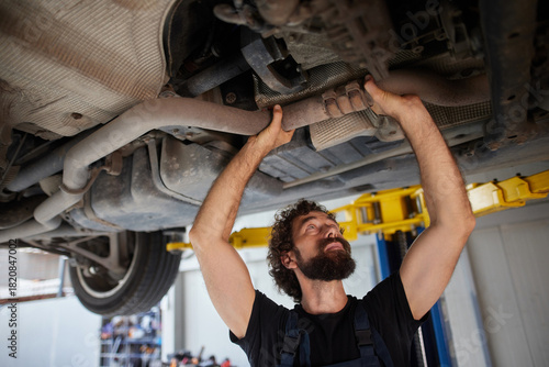 A skilled car mechanic works diligently under a raised vehicle in a busy auto repair shop. He inspects the exhaust system, demonstrating expertise and dedication to his craft.