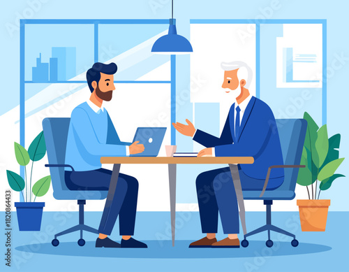 Two men in an office setting, one with a laptop, engaged in a discussion at a table. They are sitting in chairs, with a window and plants in the background.