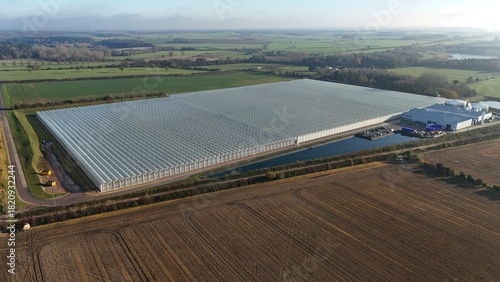 Aerial view of a vast, shimmering greenhouse complex reflecting the sky, bordered by fields, a ribbon of water, and industrial buildings, Bury Saint Edmunds, England, United Kingdom.