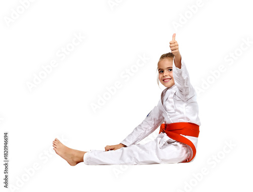 A karate girl sits on the floor in a kimono with an orange belt and shows a thumbs up