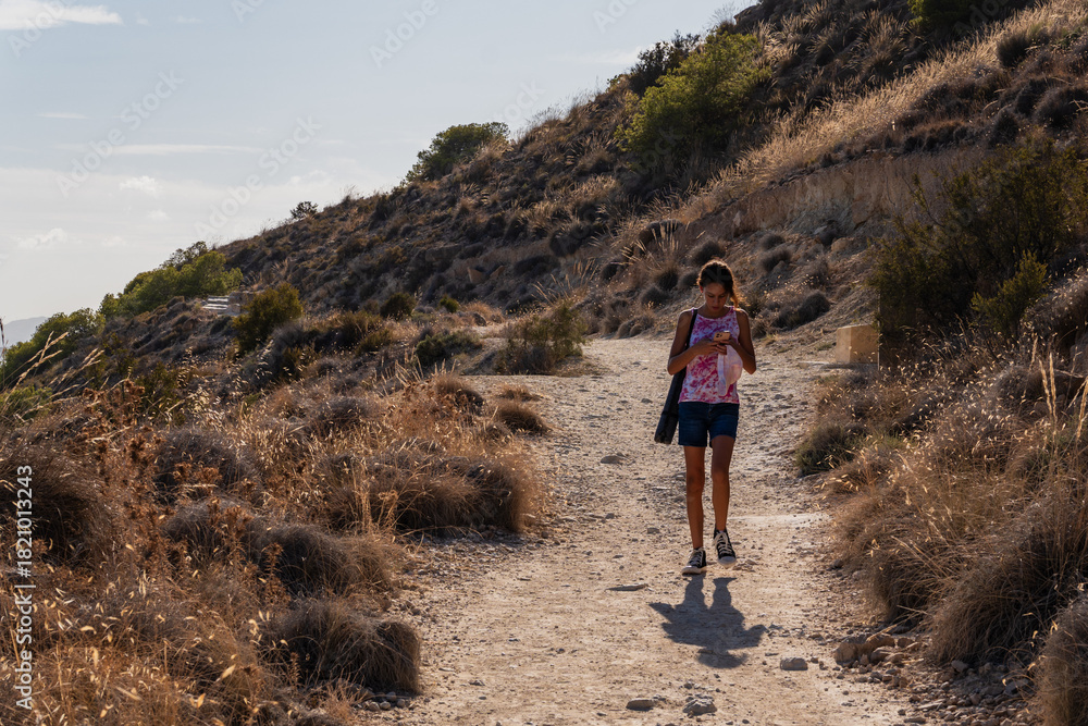 Fototapeta premium Young girl walking on a natural hiking trail with rock pavements while looking at her mobile phone. Technology and nature contrast.