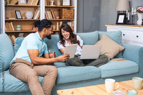Smiling couple sits on a sofa comparing a tablet and laptop, coordinating tasks for budgeting, study, or a small business project. Real home collaboration, remote work energy, positive teamwork.