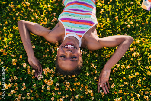 African woman laying on her back in blooming grass, smiling brightly at camera with smartphone beside, symbolizing freedom, nature, happiness, and relaxed digital lifestyle.