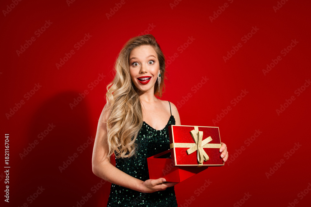 Fototapeta premium Young woman in green sequin dress presents festive red gift box with gold ribbon against red background