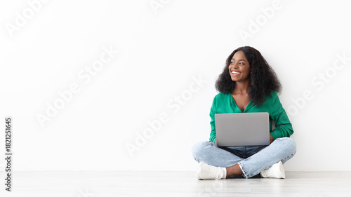 A woman sits cross-legged on the floor, smiling as she uses a laptop. She is in a bright, minimalist room with white walls, wearing a green shirt and jeans.