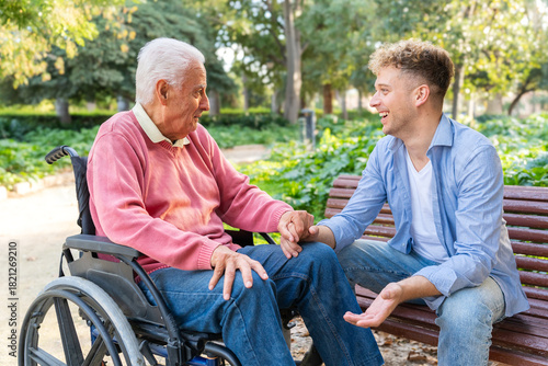 Smiling grandson holding grandfather’s hand beside grandfather seated in wheelchair on park bench