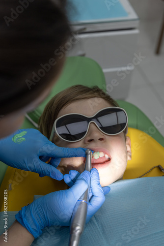 Pediatric dentist treating a boy in protective glasses, closeup of dental tools in child mouth in modern clinic.