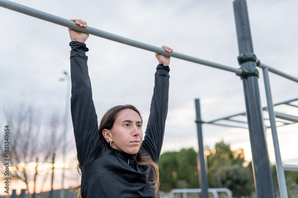 Fototapeta premium Woman performing calisthenics, hanging from a pull-up bar, training upper body strength outdoors