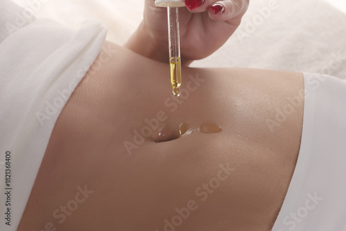 Close-up of a woman applying cosmetic oil with a dropper directly into her navel for skincare, studio shot on a white background.