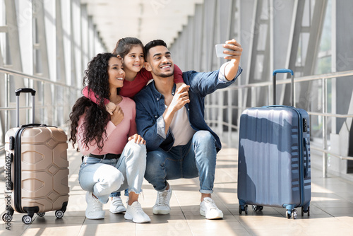 A family of three smiles as they take a selfie at the airport. They are surrounded by luggage and appear excited about their upcoming trip, capturing a joyful moment together.