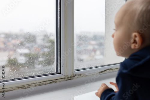 Little child looking through the window with black spots of mouldy fungus on it. Problem of condensation on the windows.