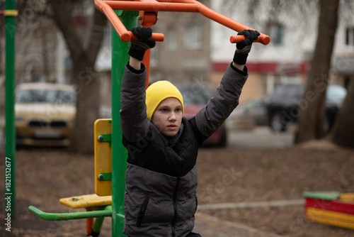 A determined child in a yellow beanie and black gloves exercises on an outdoor gym bar machines in a park, wearing a winter jacket.