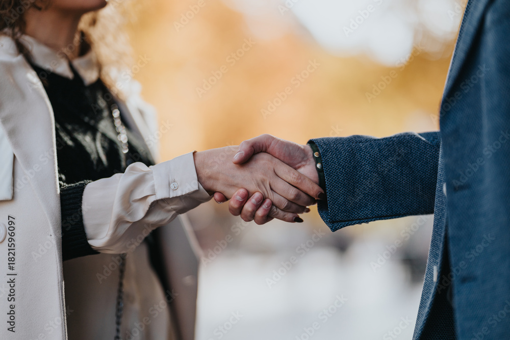 Fototapeta premium Two professionals in autumn attire exchange a handshake, symbolizing partnership and trust in a sunlit outdoor setting.