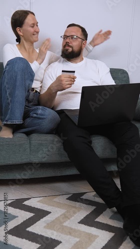 Joyful couple lounging on comfortable home sofa, sharing laptop and using credit card while enjoying seamless online shopping experience together vertical video