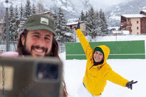 Two friends enjoy a snowy day as one takes a selfie and the other poses playfully in a bright yellow jacket. Surrounded by snow-covered trees and mountains, the