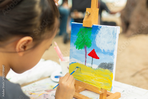 Close-up and selective focus of young girl painting a palm tree and sea with beautiful color on canvas. This shows creativity of a child's artistic hobby in a natural, outdoor beach setting.