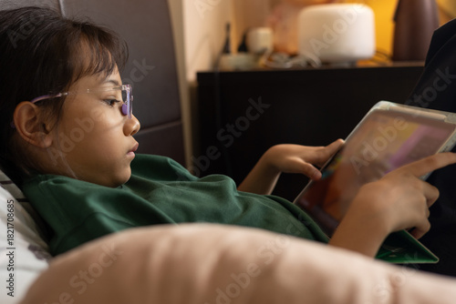 Selective focus of Asian kid girl, playing game on tablet with concentration. Concepts of children technology use, online learning, screen time, digital lifestyle, and modern childhood education.