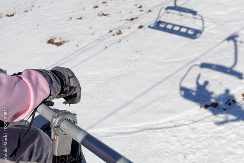 A person rides a ski lift above a snowy slope, wearing winter gloves and a pink jacket. Shadows of the lift and riders stretch across the snow, capturing a calm moment on the mountain.