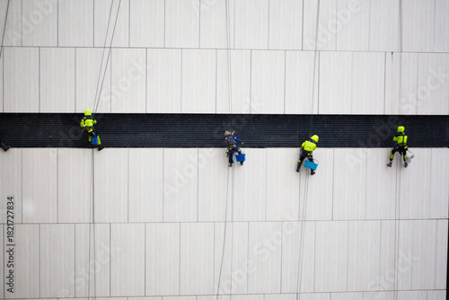 Construction crew of climbers performs dangerous task. Men in protective suits sit on seat suspended by ropes at great height. Industrial climbers work on the wall of building