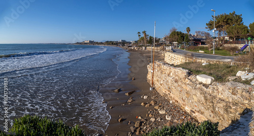 Fototapeta Naklejka Na Ścianę i Meble -  A wide panoramic view of a deserted Mediterranean beach in winter, with gentle waves arcing onto wet sand and a stone promenade lined with palms and hotels. Side, Antalya, Turkey.

