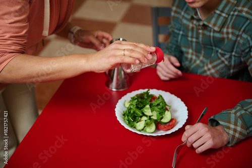 Quadro su tela Teenage Friends Sharing Salad at Red Table While Salt Is Shaken Onto Plate