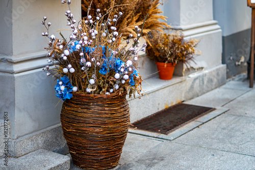 Decorative arrangement of dried plants in wicker pot featuring cotton stems and blue flowers placed beside an entrance walkway