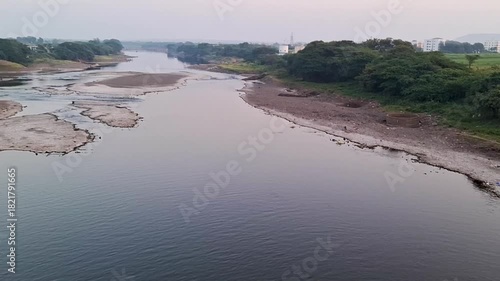A tranquil riverscape showing smooth water, exposed riverbed patches, and lush greenery along the banks. The soft pastel sky adds a peaceful atmosphere, capturing the natural beauty of a quiet river