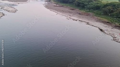 A tranquil riverscape showing smooth water, exposed riverbed patches, and lush greenery along the banks. The soft pastel sky adds a peaceful atmosphere, capturing the natural beauty of a quiet river