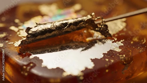 Close-up view of gold leaf being applied to a textured amber surface with iridescent reflections