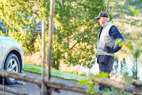 Inspection distance — mechanic stands hands-on-hips, observing parked vehicle in green yard, framed by wood fence and soft daylight.