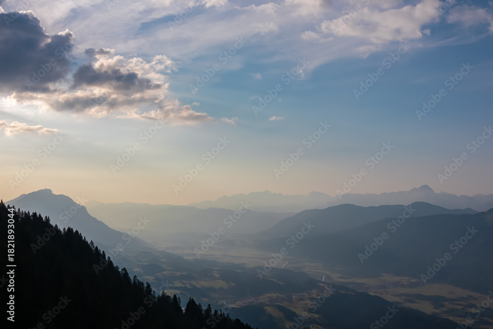 Obraz premium Sun rays stream through morning clouds, illuminating vast and hazy Gailtal valley. Serene, atmospheric view from Graslitzen, Austria, showcases layers of the Karawanks and Carnic Alps silhouettes.