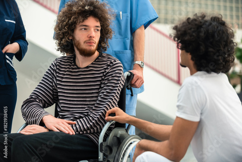 Young Man In Wheelchair Talking With Friend While Nurse Stands By In Hospital For Medical Care And Support During Recovery Process