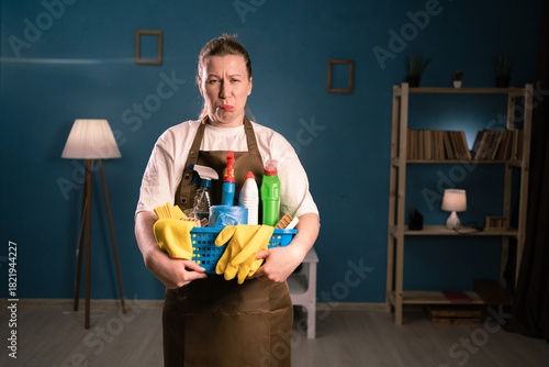 Принти на полотні Portrait of unhappy dissatisfied woman in apron holding bucket with cleaning supplies folded look camera standing in living room