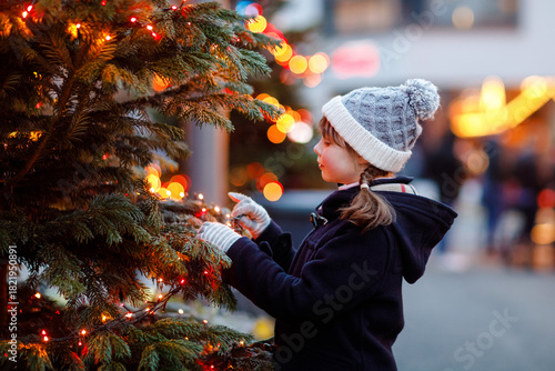 Little cute kid girl having fun on traditional Christmas market during strong snowfall. Happy child enjoying traditional family market in Germany. Schoolgirl standing by illuminated xmas tree.