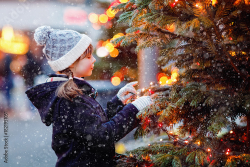 Little cute kid girl having fun on traditional Christmas market during strong snowfall. Happy child enjoying traditional family market in Germany. Schoolgirl standing by illuminated xmas tree.