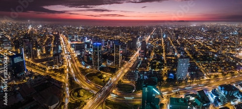 High-altitude aerial drone view of the vast Lima cityscape at night, featuring the illuminated San Isidro financial district and endless urban sprawl under a twilight horizon.