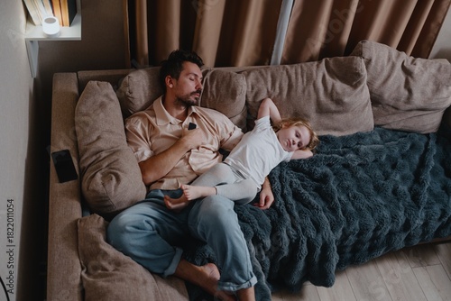A tired young father and his 4-year-old daughter watch TV together in the evening. Authentic moment of exhausted parenting and quiet family bonding