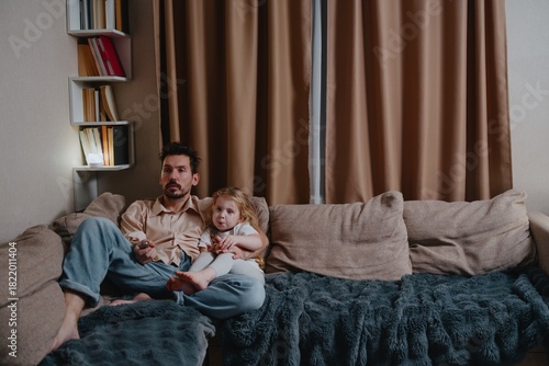 A tired young father and his 4-year-old daughter watch TV together in the evening. Authentic moment of exhausted parenting and quiet family bonding