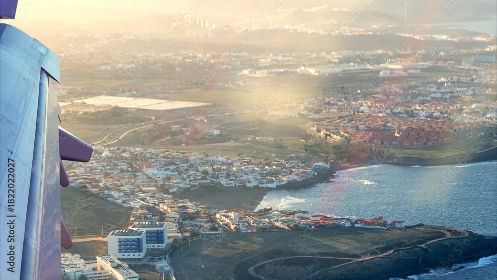 Obraz premium Airplane wing capturing an aerial view of the coastal town and residential areas of Gran Canaria, Canary Islands, during a warm sunset, symbolizing travel and holiday destinations