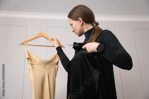 Professional stylist comparing two elegant dresses on wooden hangers, thoughtfully deciding which garment best suits her client while working in a stylish fashion atelier