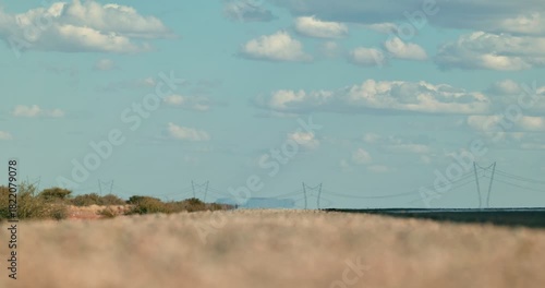 Wide shot of a desert road shimmering in the scorching heat, small clouds in a blue sky, with a distant power line and mirage-like reflection of an oncoming car on the horizon.