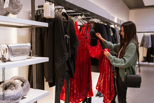 Young brunette woman choosing a red sequin dress during festive Christmas shopping, exploring holiday outfits and sparkling party looks in a fashion store
