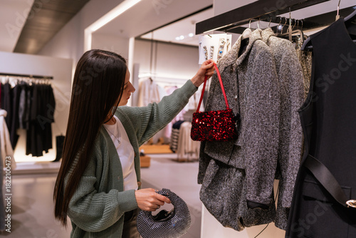 Young brunette woman shopping for Christmas season accessories, comparing glitter bags and matching them with a festive tweed outfit in a fashion store