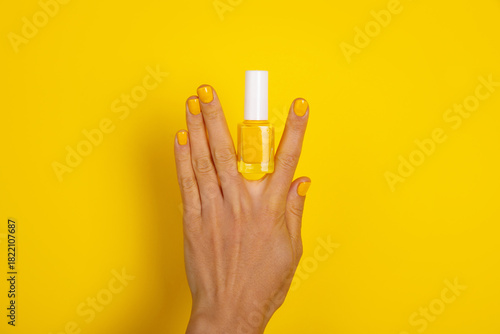 Female hand with yellow polished nails holding a yellow nail polish bottle against a bright yellow background. Modern, vibrant beauty concept.